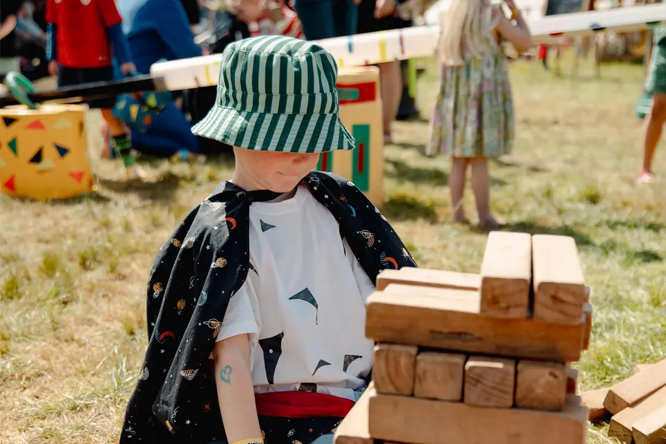 a boy in a bucket hat and cape builds a tower as tall as him with wooden blocks