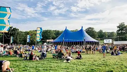 Groups of people are sat on the grass enjoying the sun in fron of the Far Out Stage, a blue circus style tent