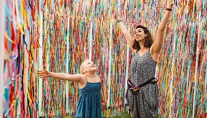 A girl and woman stand happily with their arms outstretched in a cube full of colourful ribbons