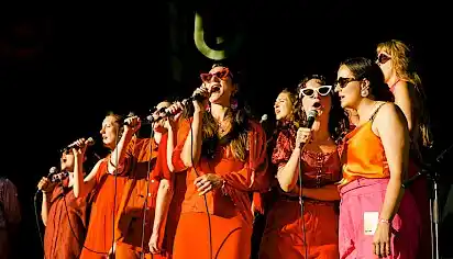A group of women dressed in orange sing into microphones, some are wearing sunglasses, the sun is shining on them