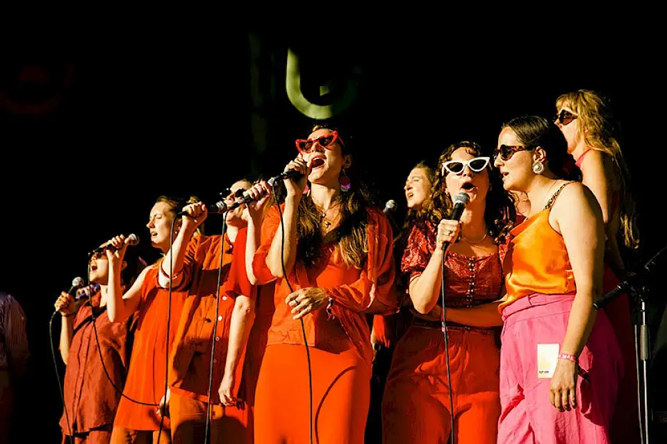 A group of women dressed in orange sing into microphones, some are wearing sunglasses, the sun is shining on them