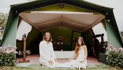 A couple dressed in white sit outside a green tent, there are gongs inside and flowers on either side of the tent
