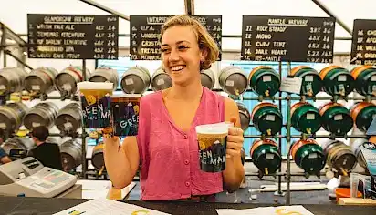 A bartender smiles as she hands three pints over the counter, behind her are many barrels of beer