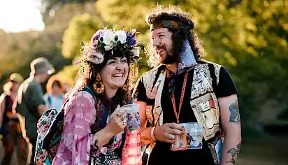 A man with a bandana and woman in a flower crown laugh at one another whilst carrying their drinks