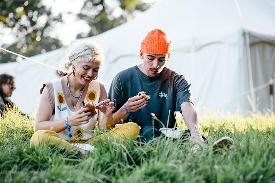 A man and woman sit amongst the grass eating their food