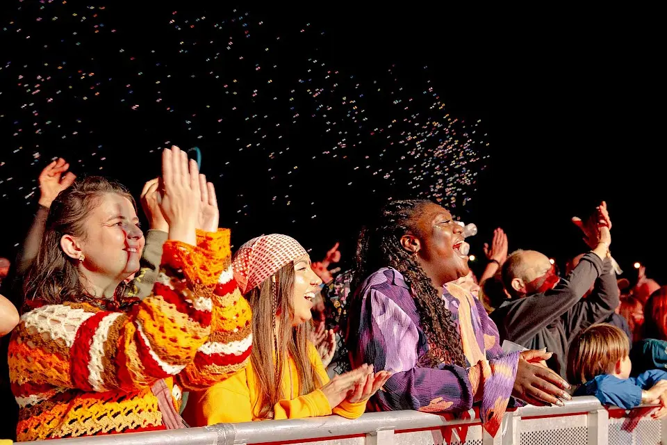 The front row of the crowd at night time, they are all dressed in bright colours, there are bubbles in the background