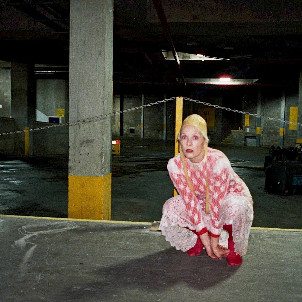 Cate Le Bon is crouching in a car park in a pink dress and yellow bonnet