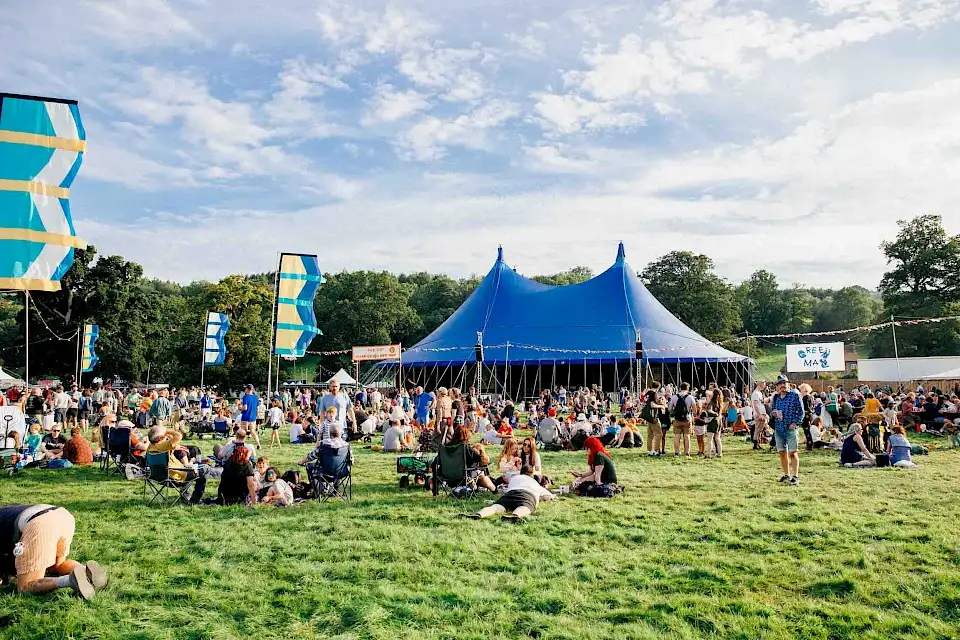Groups of people are sat on the grass enjoying the sun in fron of the Far Out Stage, a blue circus style tent