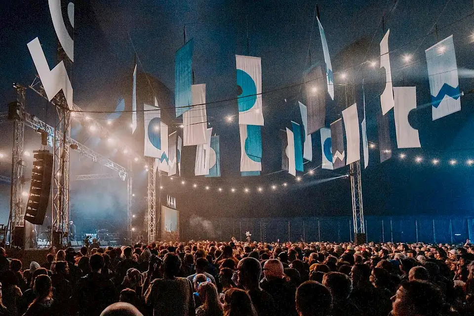 The inside of the Far Out tent, a large crowd of people all facing the stage, hanging from the ceiling are large banners with blue and white symbols.