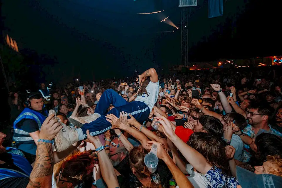 A man crowd surfs at the front of the stage whilst singing into a microphone, the audience lift him up and reach towards him.