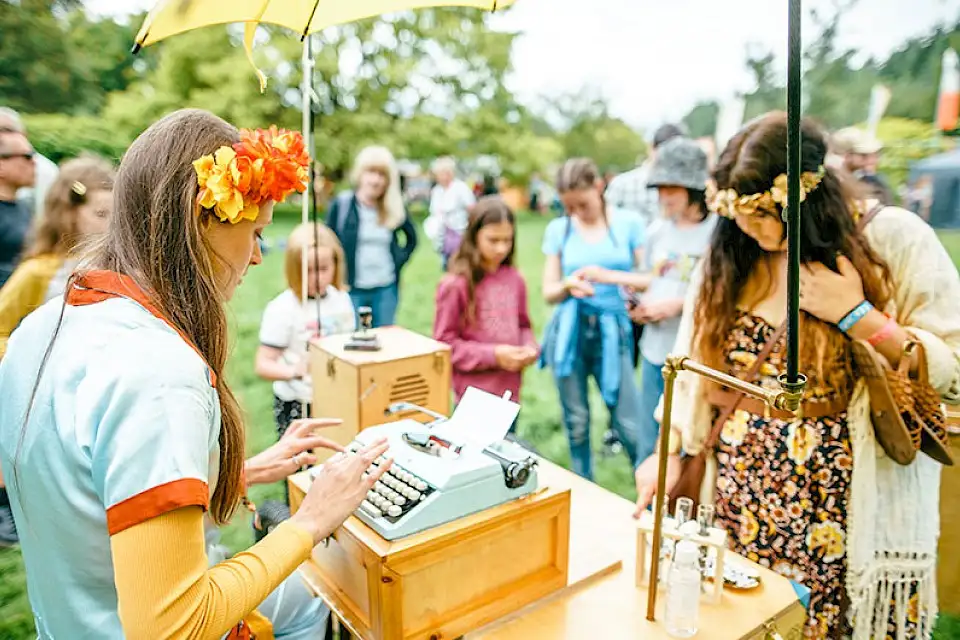 A woman with orange flowers in her hair sits and writes at a pale blue typewriter