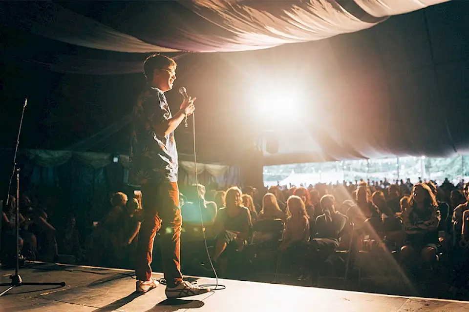 A performer is stood on stage with a mcirophone in hand as a packed tent watch on