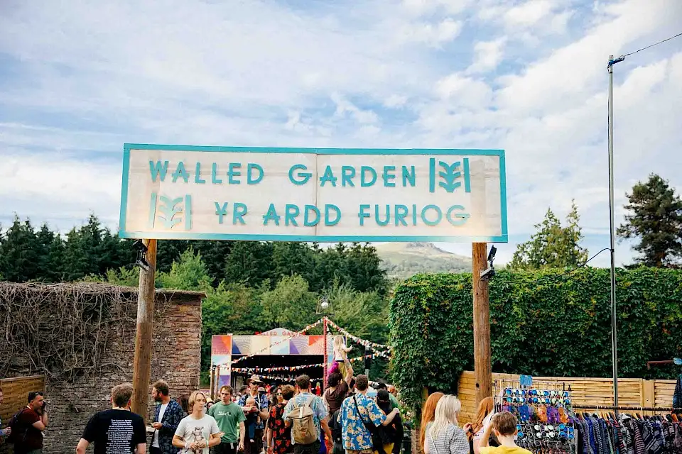 The enterance to the Walled Garden, a large sign over the walkway reading 'Walled Garden - Yr Ardd Furiog', you can see the Walled Garden stage in the background