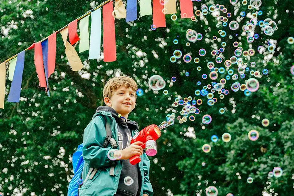 A boy stands beneath bunting with a bubble gun, he is surround by trees and bubbles