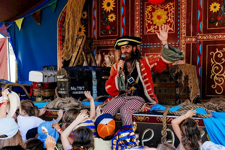 A pirate sits on stage infront of a painted backdrop of flowers and patterns as children raise gather around and raise their hands