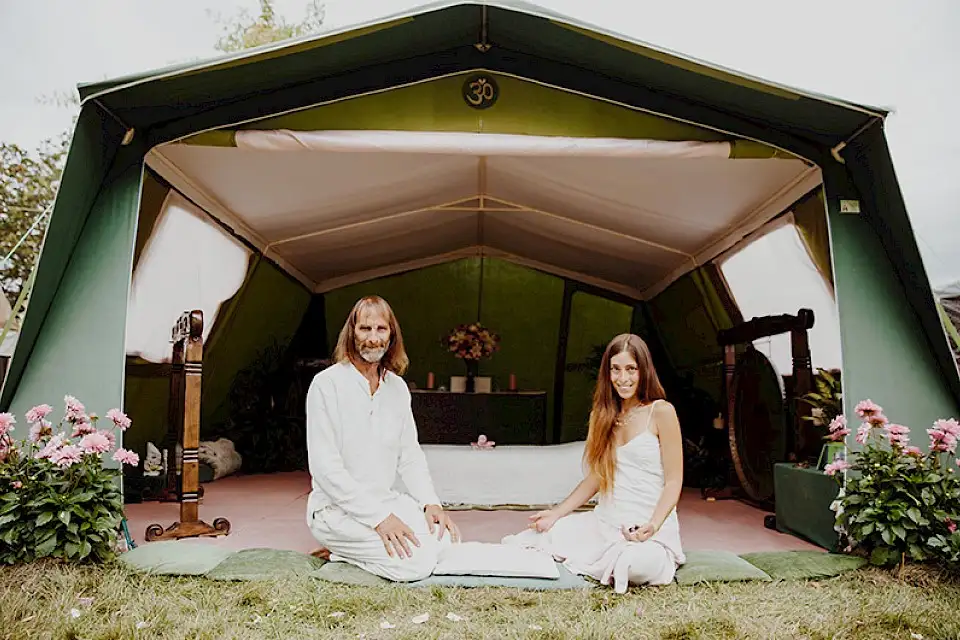 A couple dressed in white sit outside a green tent, there are gongs inside and flowers on either side of the tent