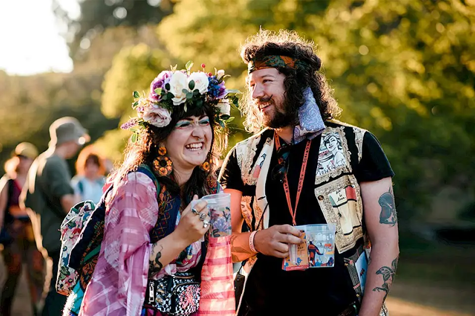 A man with a bandana and woman in a flower crown laugh at one another whilst carrying their drinks