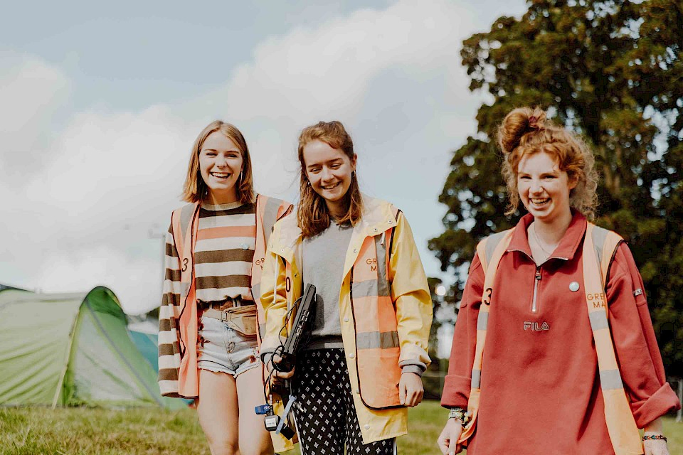 Three students with high-vis jackets and radios laugh as they walk through the festival campsite