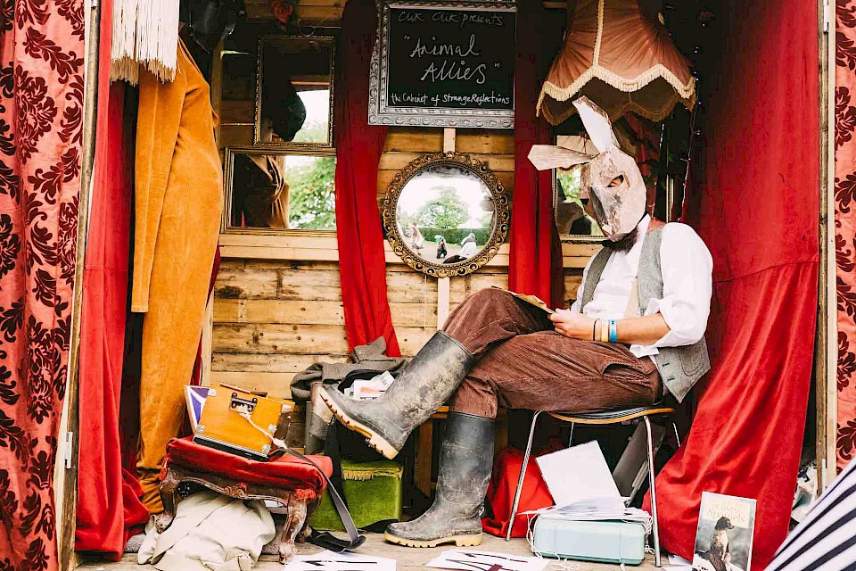 A man in a rabbit mask and wellies sits in a shed decorated with mirrors and picture frames